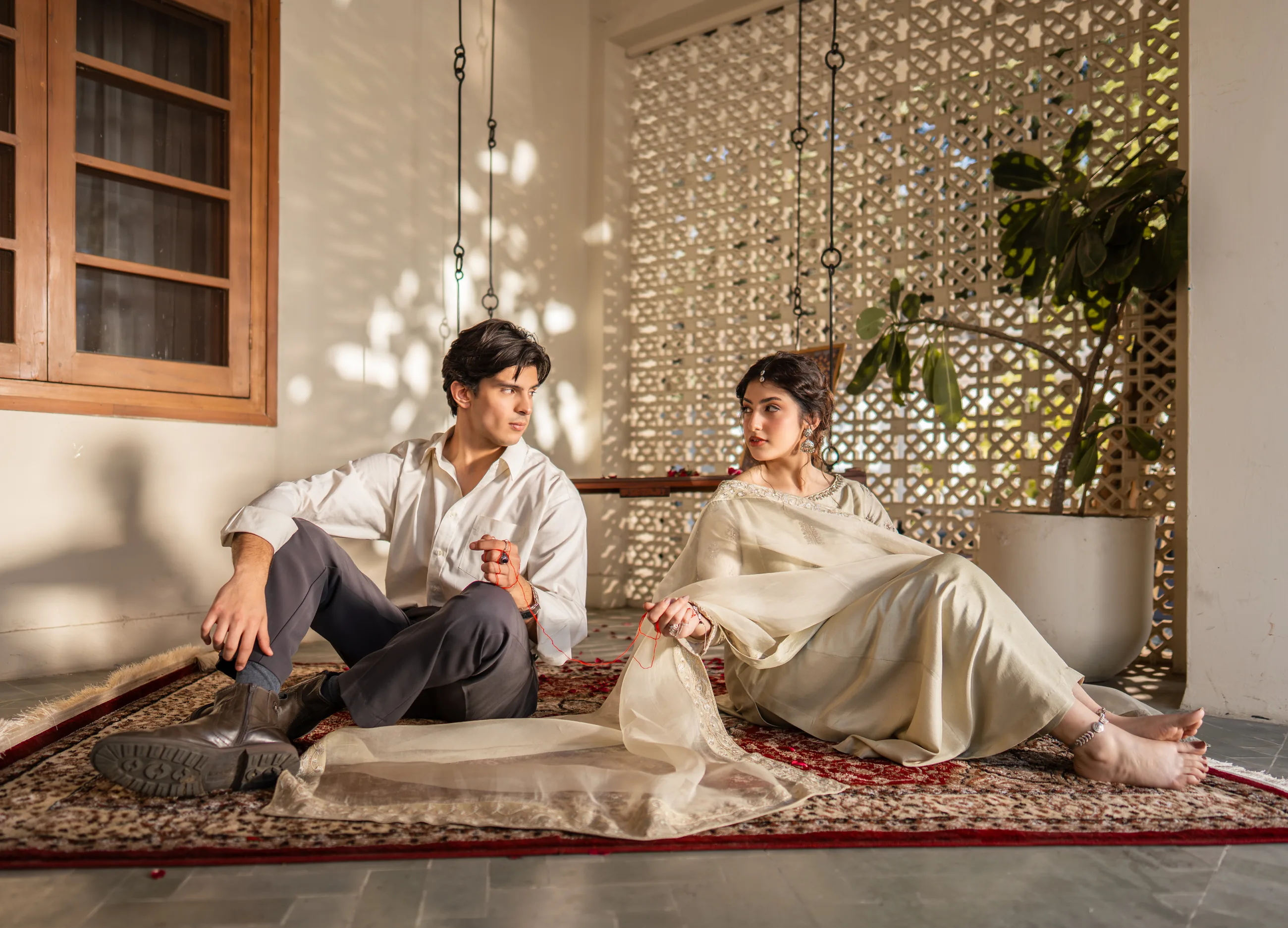 Lahore — The Ripening — a woman and a man seated together on a rug in a sunlit, jaali-screened interior, a red thread resting between them.