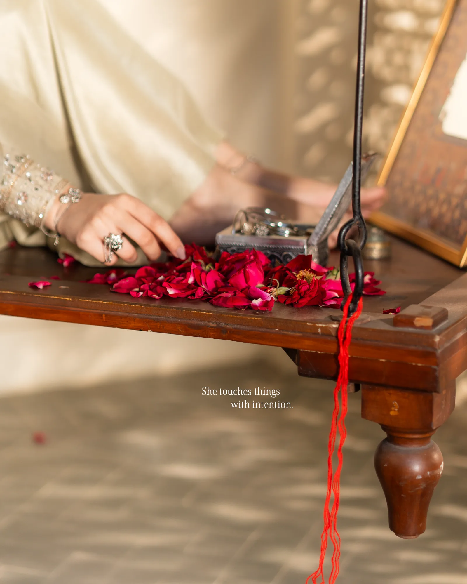 Her hand placing rose petals on a low wooden table, a red thread trailing down the table leg.