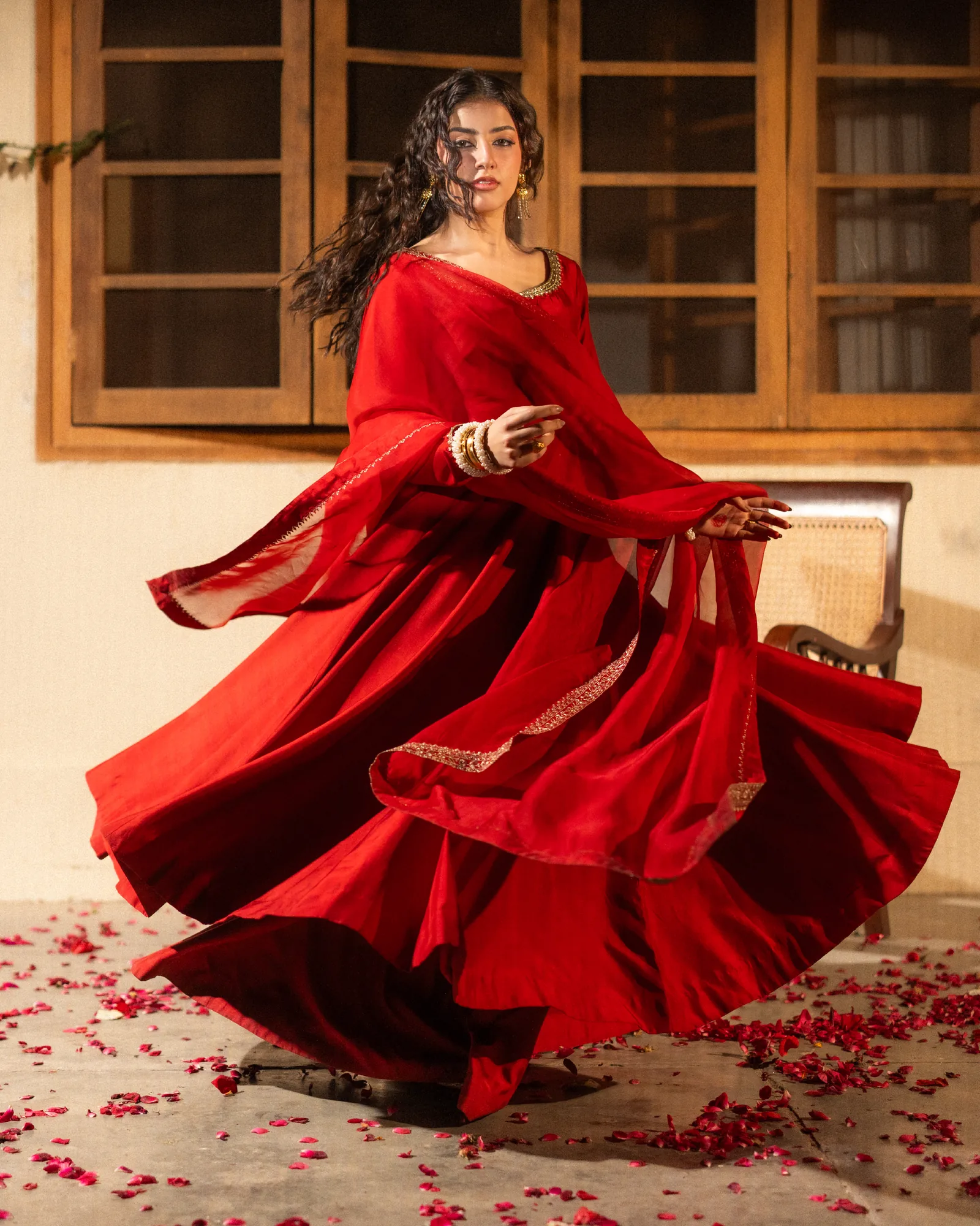 A woman turning in a flowing red peshwas, the skirt sweeping over rose petals — the bloom.