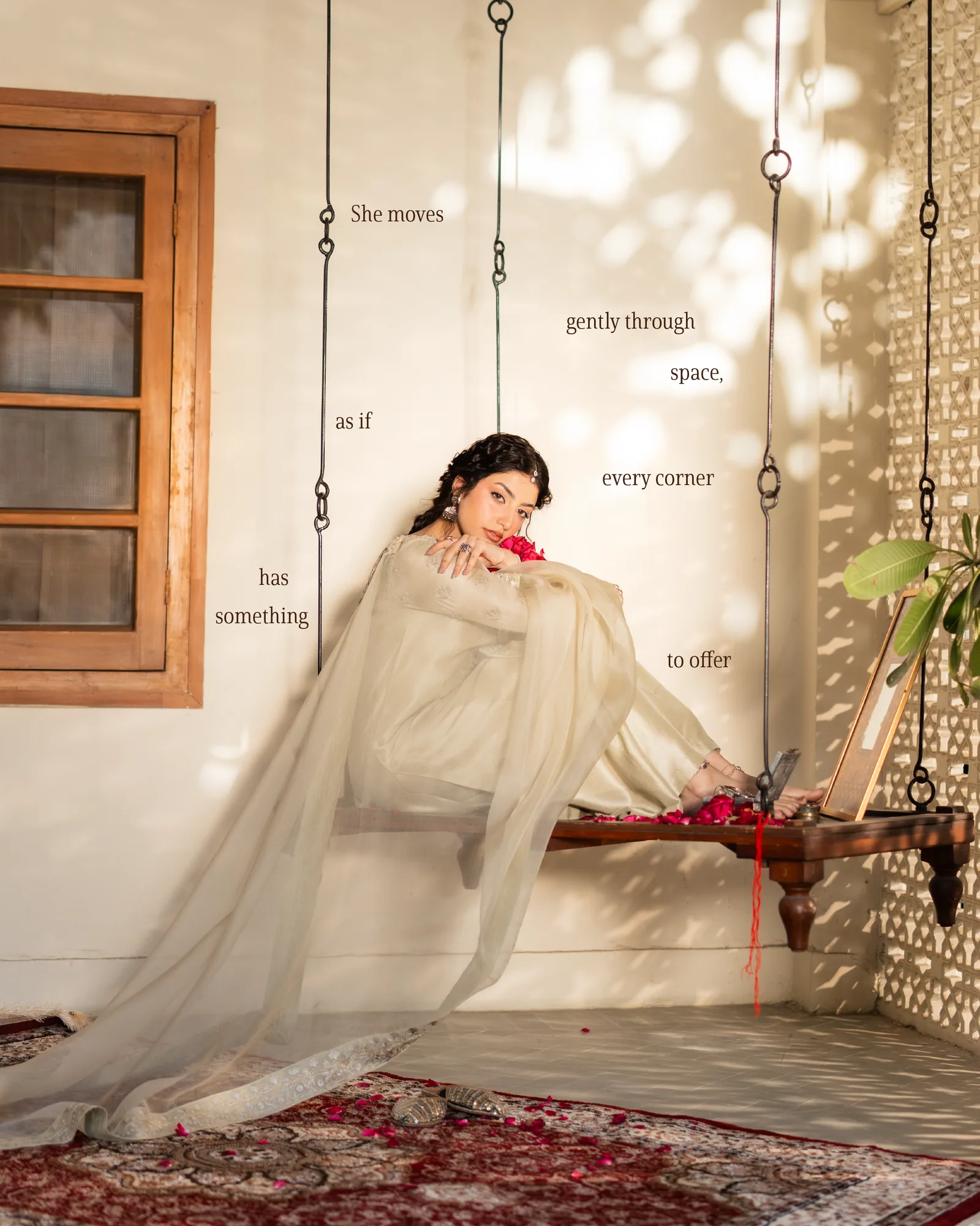 A woman in cream on a wooden swing against a jaali screen, dappled light around her, a red thread trailing to petals below.
