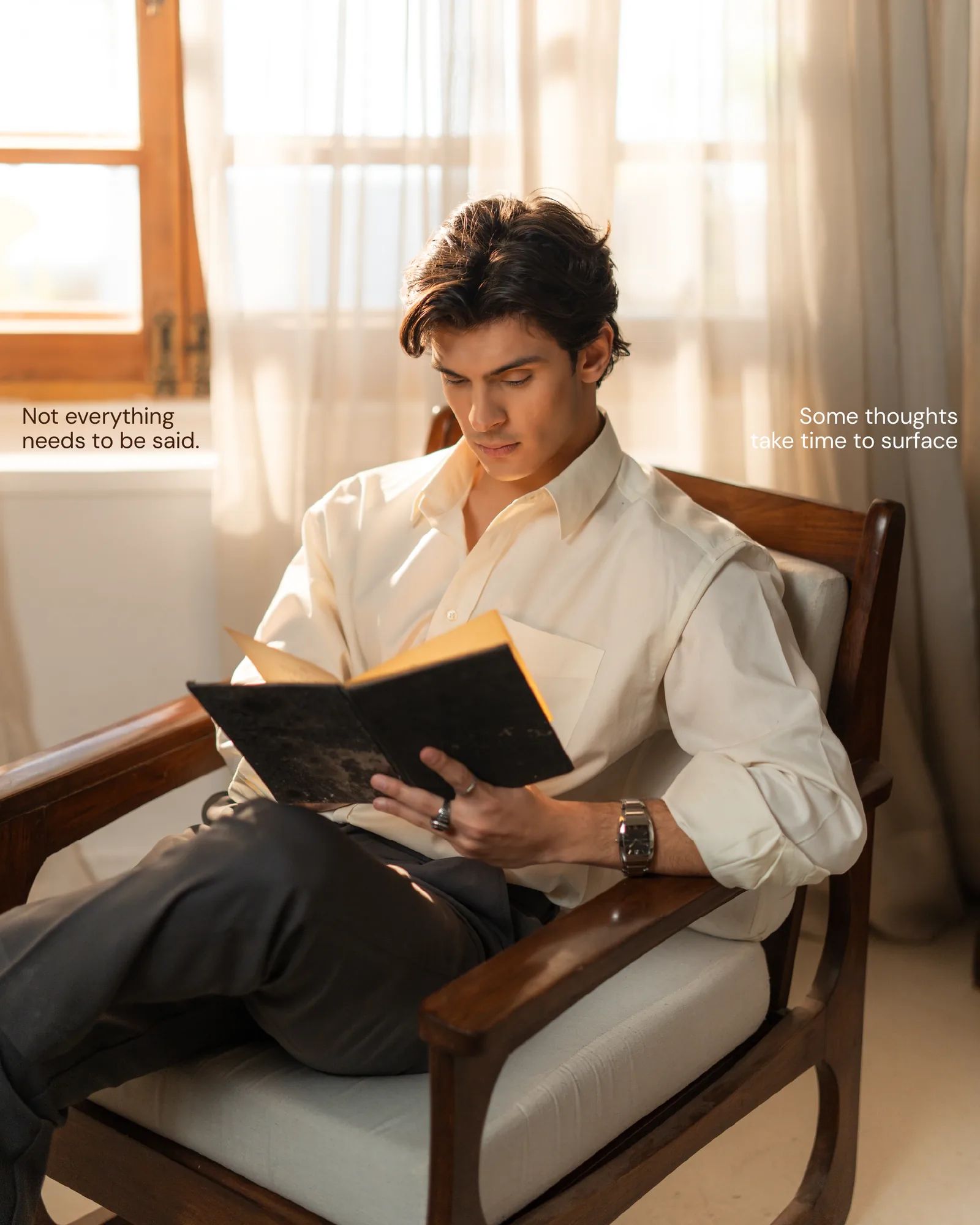 A man in a white shirt seated in a wooden chair, reading in warm window light.