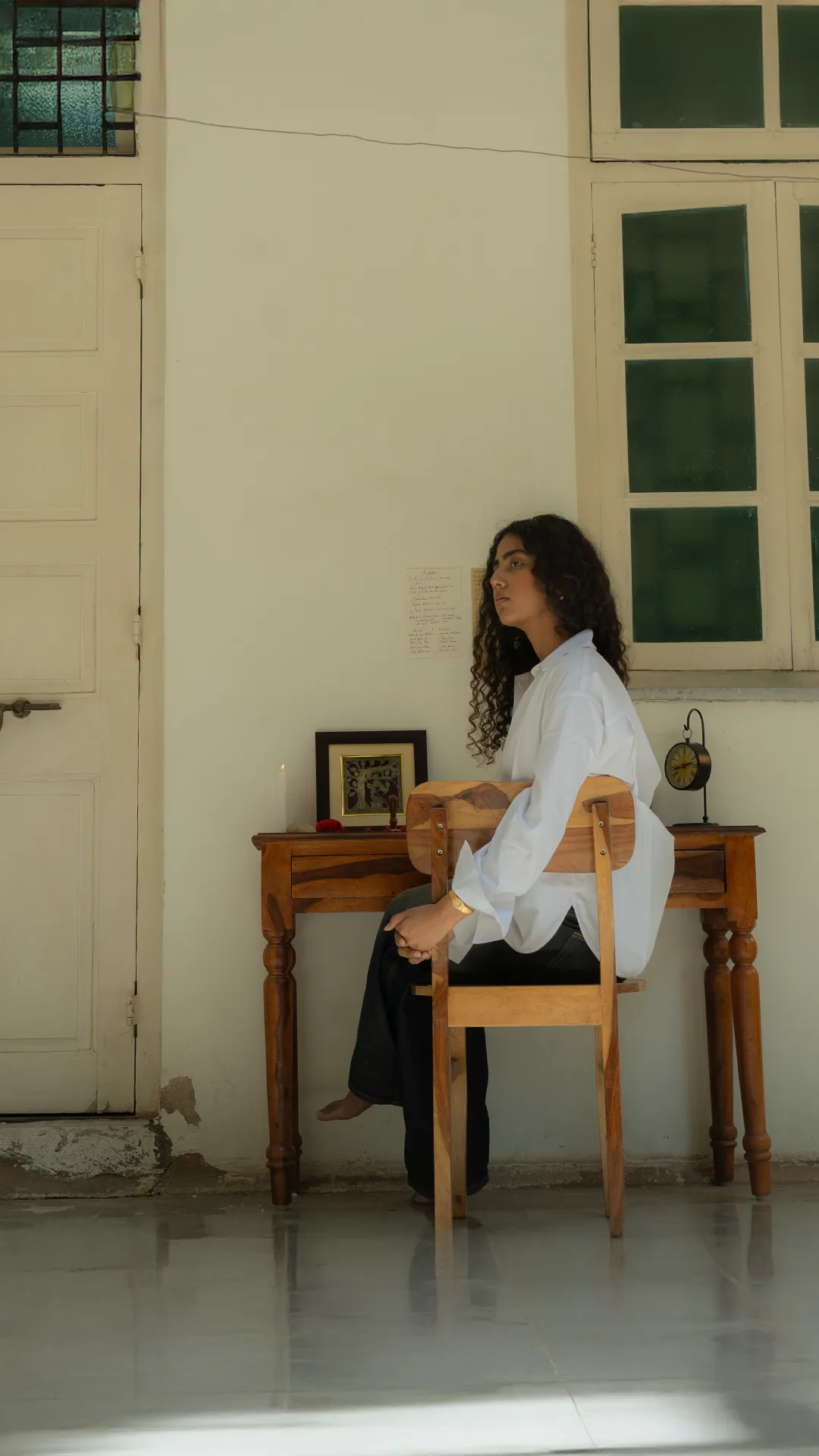 Rea seated at a wooden writing desk against a white wall and a green-paned window, in an oversized white shirt — a candle, a framed picture and a handwritten page pinned above the desk.